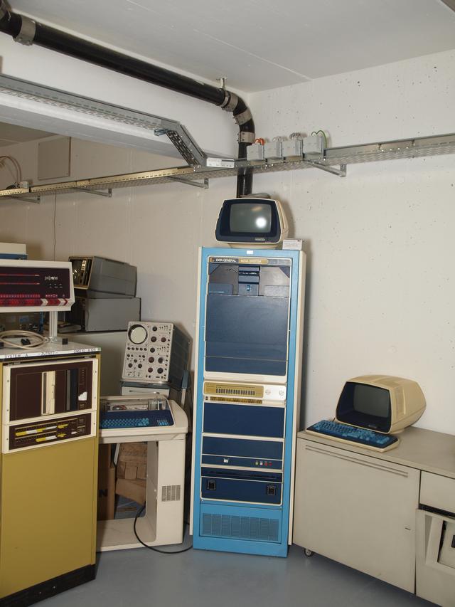 a Nova rack in the center with a terminal on the top, PDP-11/35 control panel to the left on top of some other smaller rack a Data General branded teletype terminal in betwen the two and an old large ociloscope behine. On the right a table with a glass terminal with the Data General colors.